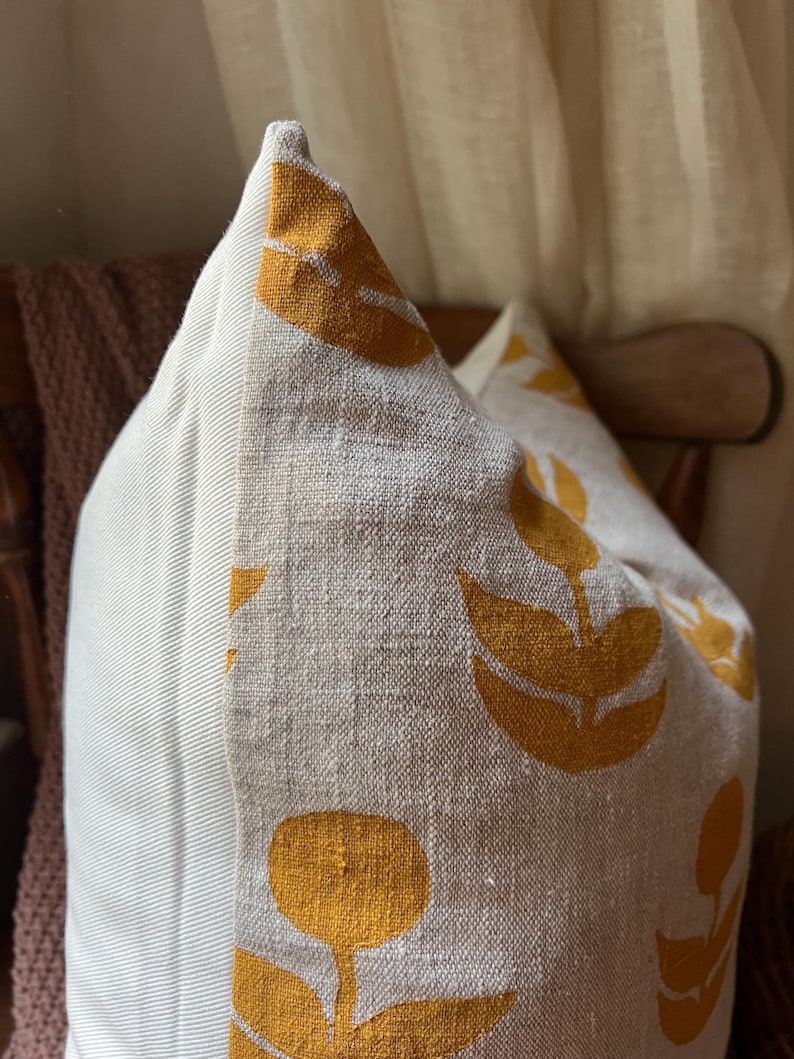 Puede incluir: Un coj&iacute;n decorativo con tela de lino beige y un patr&oacute;n repetido de dise&ntilde;os florales de color amarillo mostaza. El coj&iacute;n tiene un panel lateral a rayas y est&aacute; colocado en una silla de madera.
