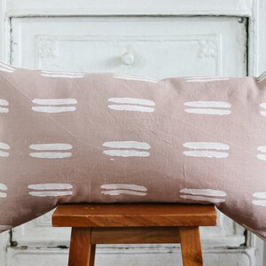 May include: A rectangular decorative pillow with a blush pink background and a repeating pattern of white horizontal brushstrokes. The pillow is resting on a small wooden stool.