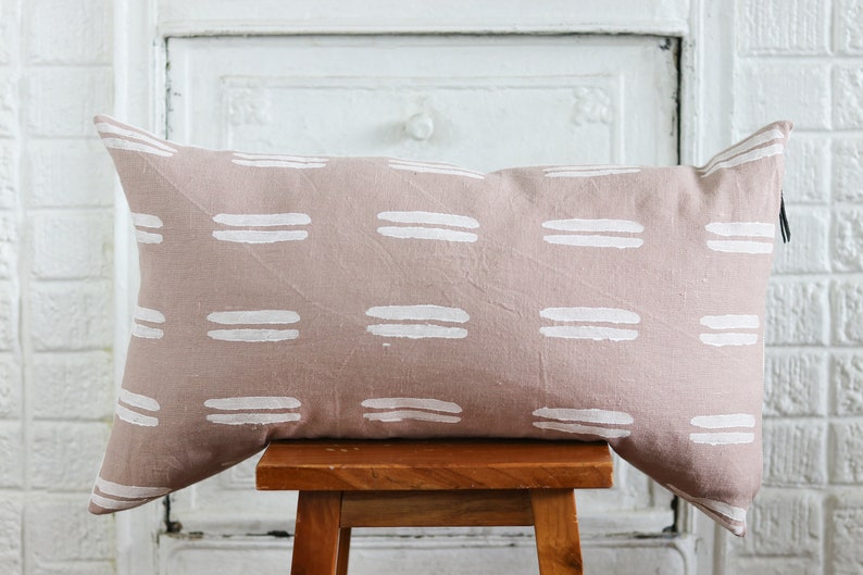 May include: A rectangular decorative pillow with a blush pink background and a repeating pattern of white horizontal brushstrokes. The pillow is resting on a small wooden stool.