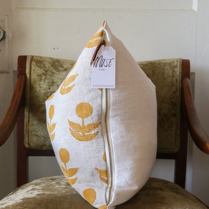 Puede incluir: Un coj&iacute;n decorativo con un estampado floral amarillo mostaza sobre fondo blanco. Tiene una cremallera blanca y una etiqueta que dice "MUSE textiles". El coj&iacute;n est&aacute; sobre una silla de terciopelo verde. Medidas en cent&iacute;metros.
