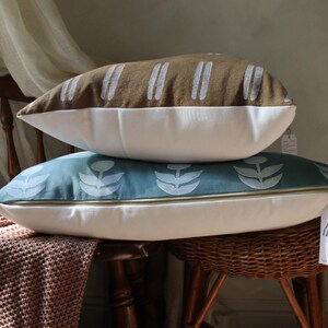May include: Two decorative pillows stacked on a wooden chair. The top pillow is brown with white vertical stripes. The bottom pillow is teal with a white floral pattern. Both pillows have white bottoms and gold zippers.