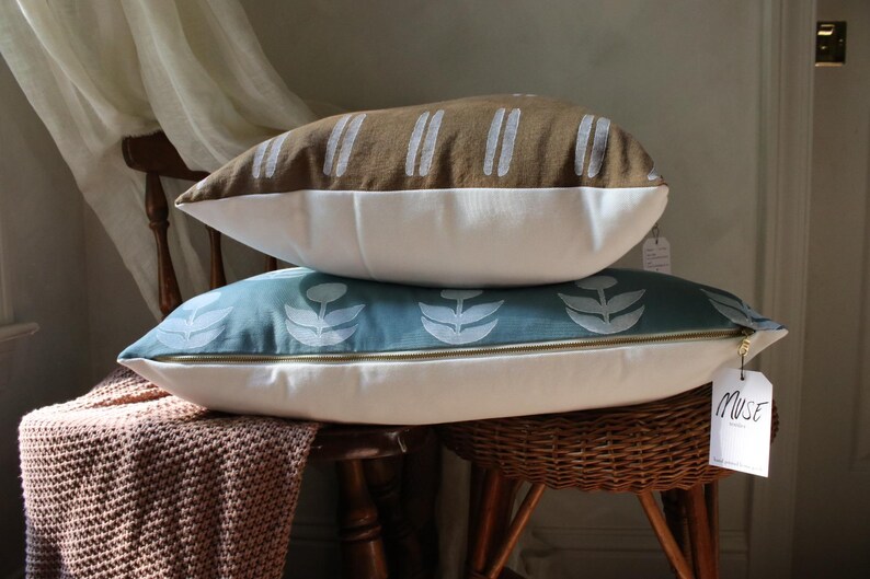 May include: Two decorative pillows stacked on a wooden chair. The top pillow is brown with white vertical stripes. The bottom pillow is teal with a white floral pattern. Both pillows have white bottoms and gold zippers.