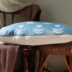 May include: A decorative pillow with a light blue floral pattern on top and a white bottom. The pillow has a gold zipper and a tag that says "Muse". The pillow is resting on a wooden chair with a woven seat.