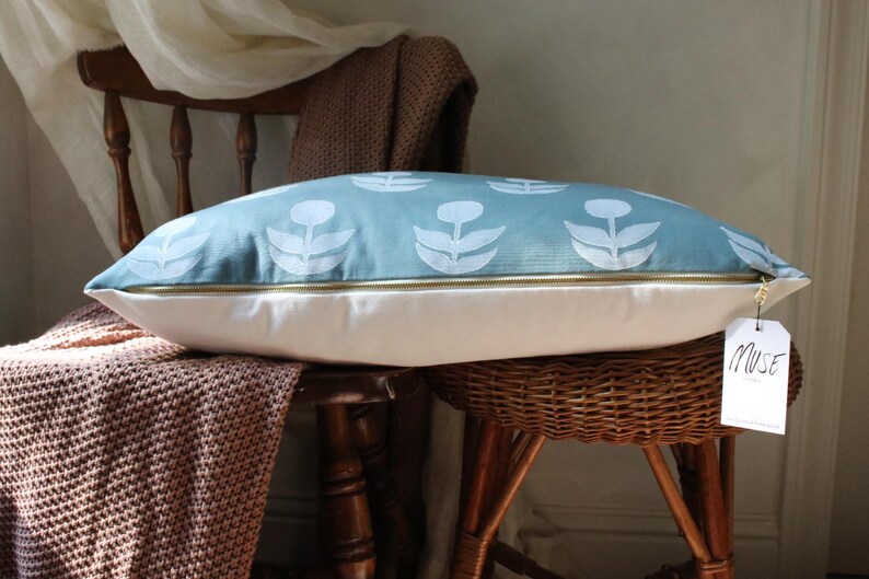 May include: A decorative pillow with a light blue floral pattern on top and a white bottom. The pillow has a gold zipper and a tag that says "Muse". The pillow is resting on a wooden chair with a woven seat.
