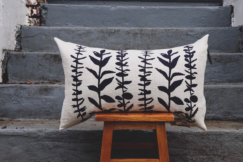 Pu&ograve; includere: Un cuscino decorativo rettangolare con sfondo color crema e motivo botanico nero ripetuto. Il cuscino &egrave; appoggiato su uno sgabello di legno, con una scala di cemento grigio sullo sfondo.