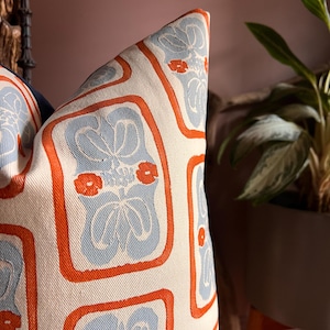 May include: Decorative pillow with a repeating pattern of light blue floral designs and small orange flowers, framed by orange borders, on a cream-colored background. The pillow is a square shape and appears to be made of a woven fabric.