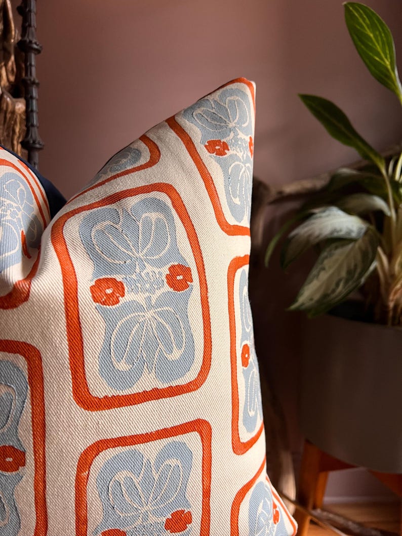 May include: Decorative pillow with a repeating pattern of light blue floral designs and small orange flowers, framed by orange borders, on a cream-colored background. The pillow is a square shape and appears to be made of a woven fabric.