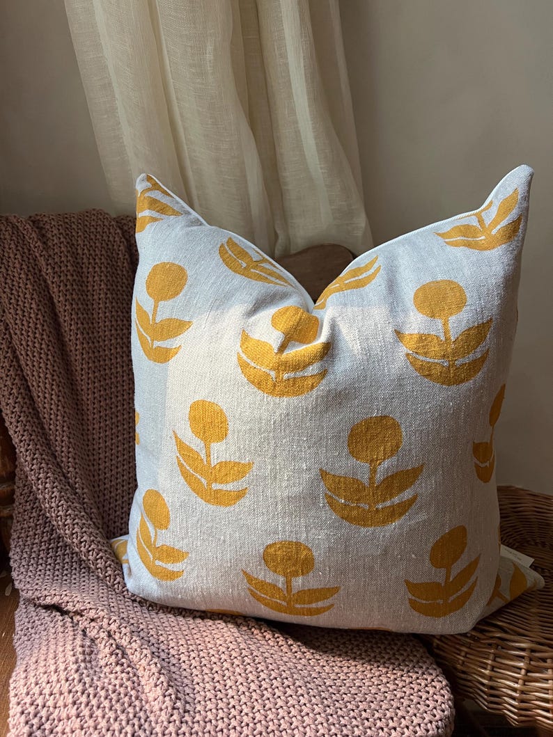 Puede incluir: Un coj&iacute;n decorativo cuadrado con un fondo gris claro y un patr&oacute;n repetido de dise&ntilde;os florales amarillos. El coj&iacute;n est&aacute; colocado sobre una manta de punto rosa y una silla de mimbre.