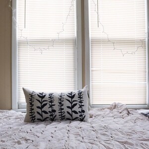 Pu&ograve; includere: Un cuscino rettangolare con un design botanico nero poggia su un letto coperto da una trapunta fantasia. Il design del cuscino presenta steli verticali con foglie. Due finestre con persiane sono sullo sfondo.