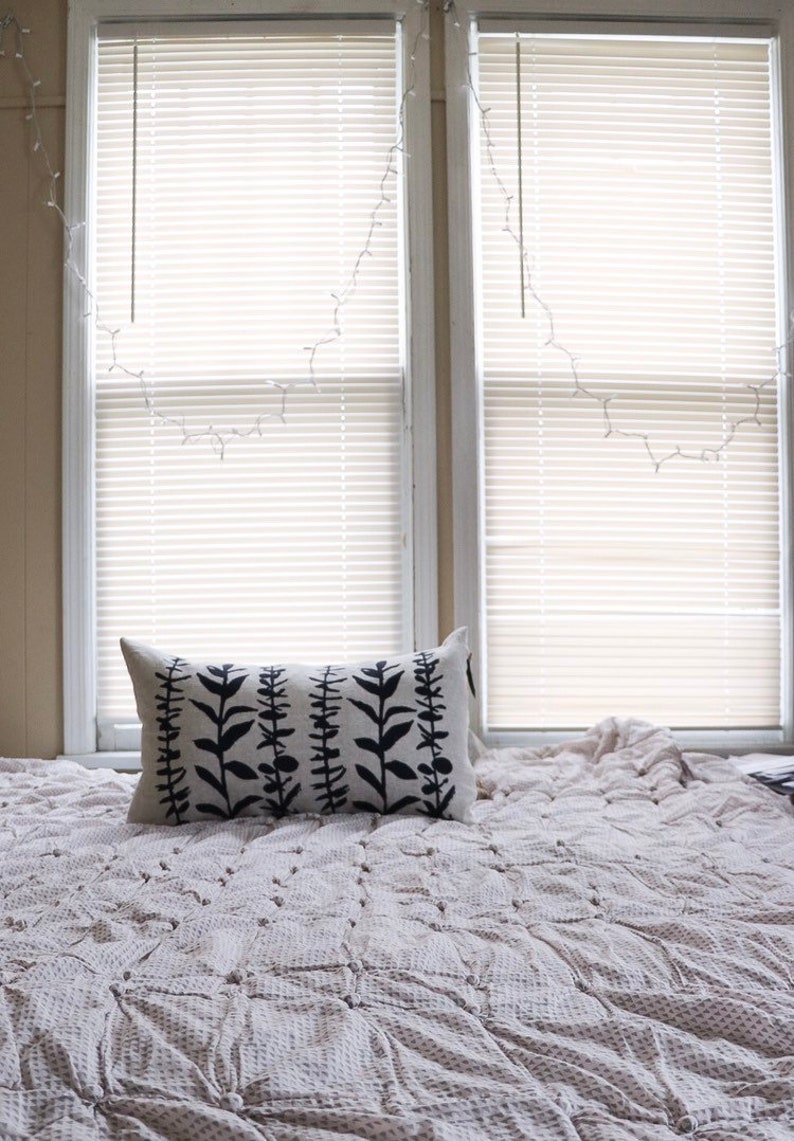 Pu&ograve; includere: Un cuscino rettangolare con un design botanico nero poggia su un letto coperto da una trapunta fantasia. Il design del cuscino presenta steli verticali con foglie. Due finestre con persiane sono sullo sfondo.