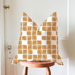 Puede incluir: Un cojín decorativo con un patrón geométrico. El cojín presenta un diseño repetitivo de formas rectangulares doradas sobre un fondo blanco. Se coloca sobre un taburete de madera frente a una puerta blanca. Medidas: aproximadamente 40 cm x 40 cm.