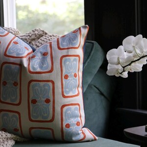 May include: A square throw pillow featuring a cream-colored fabric with a repeating pattern of light blue floral motifs enclosed in orange rectangular borders. The pillow has a dark blue side panel and is positioned against a dark green sofa.