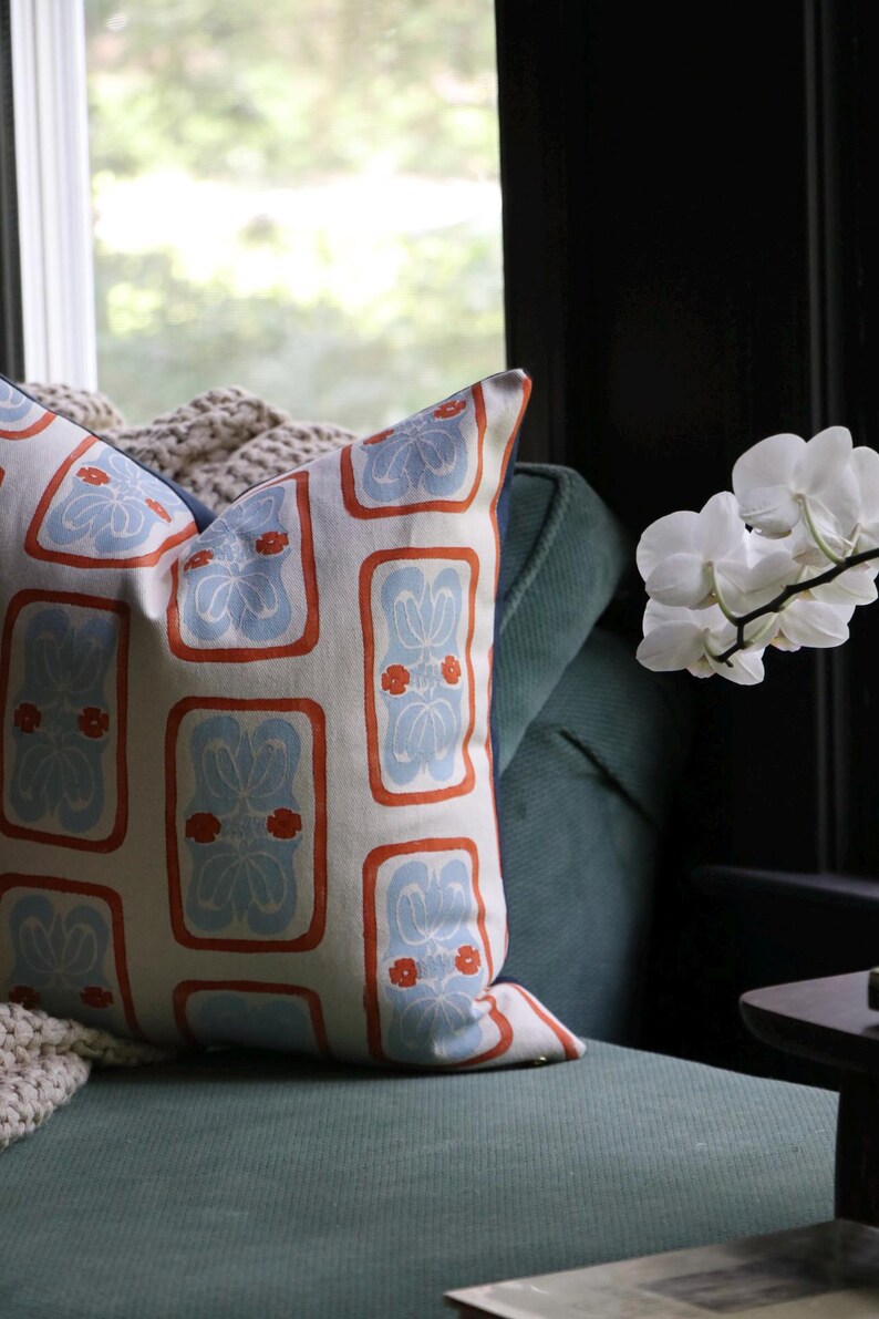 May include: A square throw pillow featuring a cream-colored fabric with a repeating pattern of light blue floral motifs enclosed in orange rectangular borders. The pillow has a dark blue side panel and is positioned against a dark green sofa.
