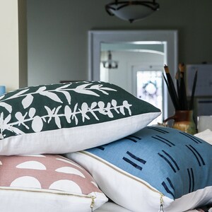 Puede incluir: Tres almohadas decorativas con diferentes patrones. La almohada superior tiene un fondo verde con dise&ntilde;os de hojas blancas. La almohada del medio tiene un fondo rosa con semic&iacute;rculos blancos. La almohada inferior tiene un fondo azul con rayas blancas. Las almohadas est&aacute;n apiladas una encima de la otra y tienen un respaldo de tela blanca. Una superficie marr&oacute;n es visible debajo de las almohadas. Una etiqueta blanca con el texto "Muse Textiles Hand printed home goods" est&aacute; unida a la almohada inferior.