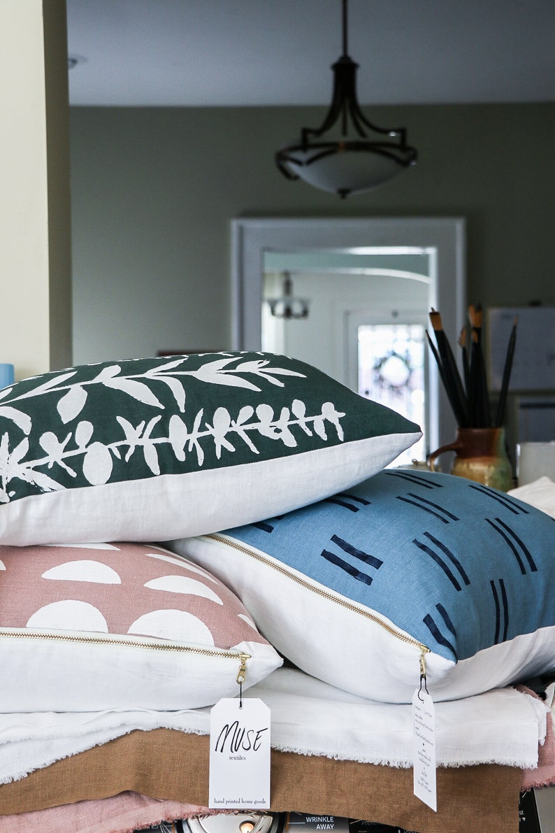 Puede incluir: Tres almohadas decorativas con diferentes patrones. La almohada superior tiene un fondo verde con dise&ntilde;os de hojas blancas. La almohada del medio tiene un fondo rosa con semic&iacute;rculos blancos. La almohada inferior tiene un fondo azul con rayas blancas. Las almohadas est&aacute;n apiladas una encima de la otra y tienen un respaldo de tela blanca. Una superficie marr&oacute;n es visible debajo de las almohadas. Una etiqueta blanca con el texto "Muse Textiles Hand printed home goods" est&aacute; unida a la almohada inferior.
