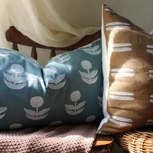May include: Two decorative throw pillows. One is teal with a white floral pattern. The other is brown with a white horizontal stripe design. The pillows are on a wooden chair with a woven seat and a brown blanket.