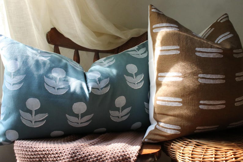 May include: Two decorative throw pillows. One is teal with a white floral pattern. The other is brown with a white horizontal stripe design. The pillows are on a wooden chair with a woven seat and a brown blanket.