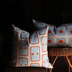 May include: Two square throw pillows featuring a repeating pattern of light blue floral motifs enclosed in orange rectangular borders on a cream background. The pillows are displayed on wooden and wicker furniture pieces in a dimly lit setting.