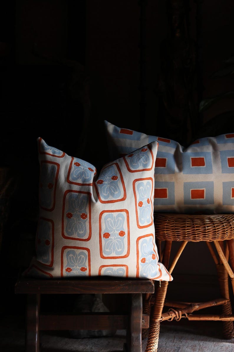 May include: Two square throw pillows featuring a repeating pattern of light blue floral motifs enclosed in orange rectangular borders on a cream background. The pillows are displayed on wooden and wicker furniture pieces in a dimly lit setting.