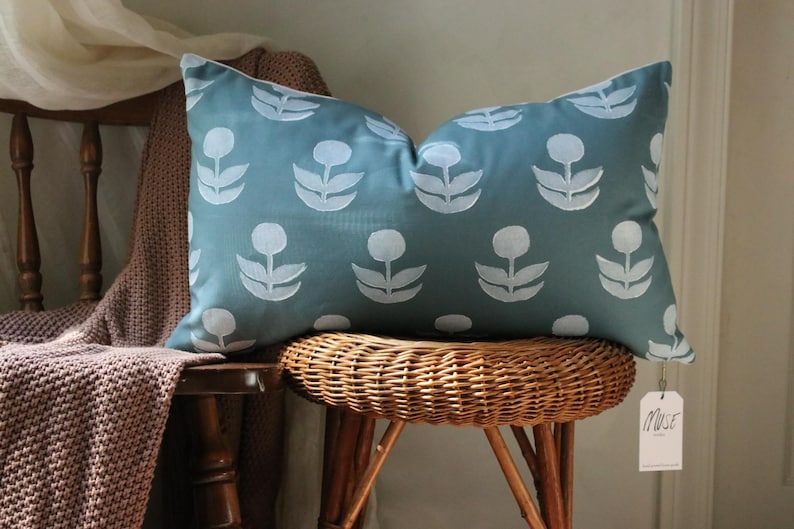 May include: A teal decorative pillow with a white floral pattern rests on a wicker chair. The pillow is rectangular and features a repeating design of stylized flowers. A tag that says "Muse" hangs from the pillow.