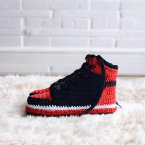 May include: Crocheted black and red high-top slippers with a white stripe. The slippers are on a white fluffy surface.