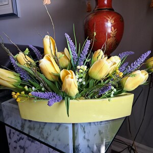 Pode incluir: Um vaso de cerâmica amarelo com forma curva contém um buquê de tulipas amarelas e lavanda roxa.