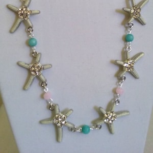 May include: A silver necklace with white and turquoise beads and five silver starfish charms. The necklace is paired with matching earrings with turquoise beads.