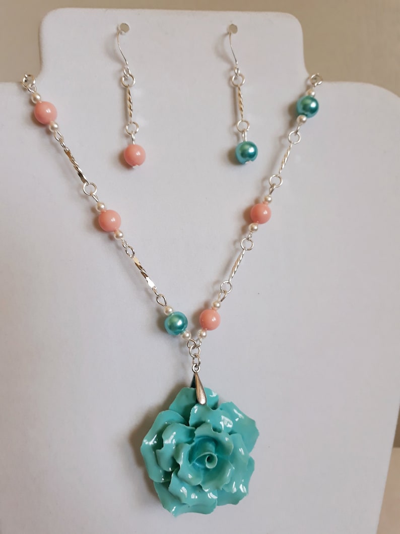 K&ouml;nnte beinhalten: Eine silberne Halskette mit einem t&uuml;rkisfarbenen Rosenanh&auml;nger und rosa und wei&szlig;en Perlen. Passende t&uuml;rkisfarbene und rosa Ohrringe sind im Lieferumfang enthalten.