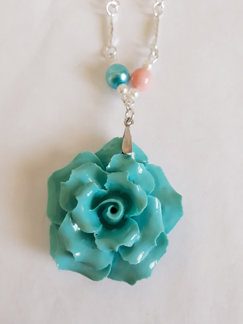 K&ouml;nnte beinhalten: Eine Halskette mit einem Anh&auml;nger in Form einer t&uuml;rkisblauen Rose, einer silbernen Kette und rosa und blauen Perlenakzenten.
