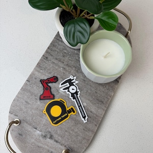 May include: A decorative marble tray with gold handles holds a small potted plant, a candle in a green-rimmed holder, and three stickers. The stickers depict a red robot arm, a black and white caliper labeled "Mechanical Engineer," and a yellow tape measure with "Cutting Twice."