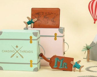 May include: A brown leather passport holder with a world map design, a green and white suitcase with the text "Chasing Threads" and a brown leather luggage tag with the text "Mr." embroidered in green thread. The items are arranged on a light yellow background with a paper airplane, a hot air balloon, and mountains in the background.