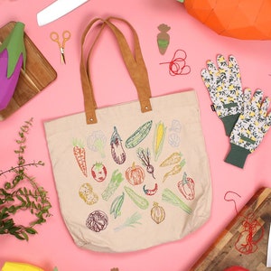 Può includere: Una borsa tote beige con verdure ricamate colorate, tra cui carote, cipolle, peperoni e melanzane. La borsa è su una superficie rosa con altri materiali per l'artigianato, come forbici, filo e carta.