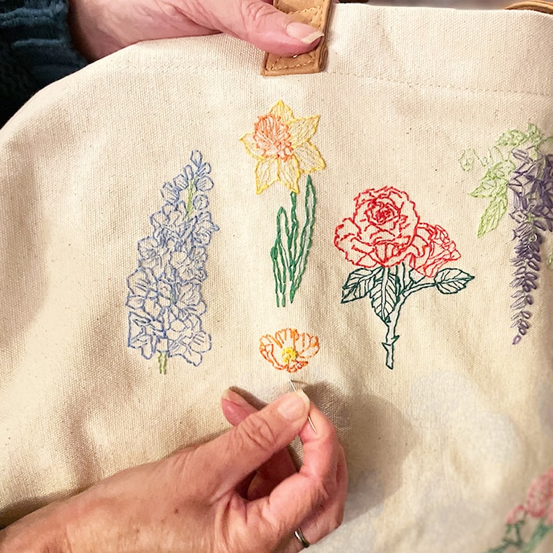 May include: A white canvas tote bag with embroidered flowers in various colors, including blue, yellow, red, and green. The flowers are arranged in a row across the bag, and the person is in the process of embroidering a small orange flower.