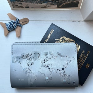 Op de afbeelding: Een grijze paspoorthouder met een gestikt wereldkaartontwerp. De houder heeft een paspoort met de tekst "Verenigd Koninkrijk van Groot-Brittannië en Noord-Ierland" en "Paspoort" zichtbaar.