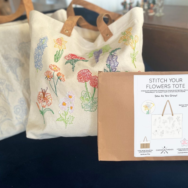 May include: A beige canvas tote bag with colorful embroidered flowers. The bag is packaged in a brown box with a white label that reads "Stitch Your Flowers Tote" and "Sew As You Grow!" with a graphic of a tote bag with embroidered flowers.