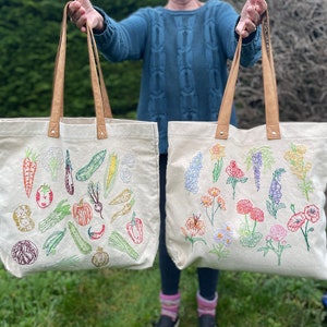 May include: Two canvas tote bags with embroidered designs. One bag features a colorful array of vegetables, while the other features a variety of flowers in different colors.
