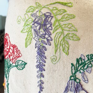 May include: Close-up of a white fabric with embroidered flowers in shades of green, purple, and red. The flowers are detailed and realistic, with delicate stitching.