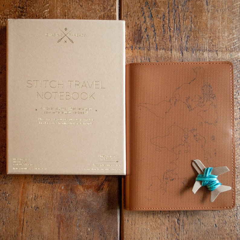 May include: A brown leather travel journal with a stitched design and a paper cover that says "Stitch Travel Notebook" in gold lettering. The paper cover also has a small illustration of a needle and thread.