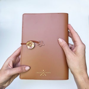 May include: A brown leather journal with a gold globe and airplane charm. The journal has the text "Chasing Threads" embossed on the front.