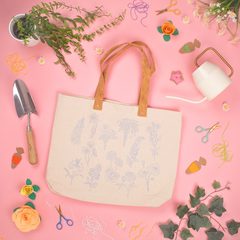 May include: A white canvas tote bag with brown leather handles and a blue floral design. The bag is surrounded by various craft supplies, including yarn, scissors, and flowers.