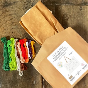 May include: A craft kit for a tote bag embroidery project. The kit includes a brown canvas tote bag, embroidery floss in various colors, and a pattern sheet with instructions. The pattern features a floral design with the text "Stitch Your Flowers Tote" and "Sew As You Grow!"