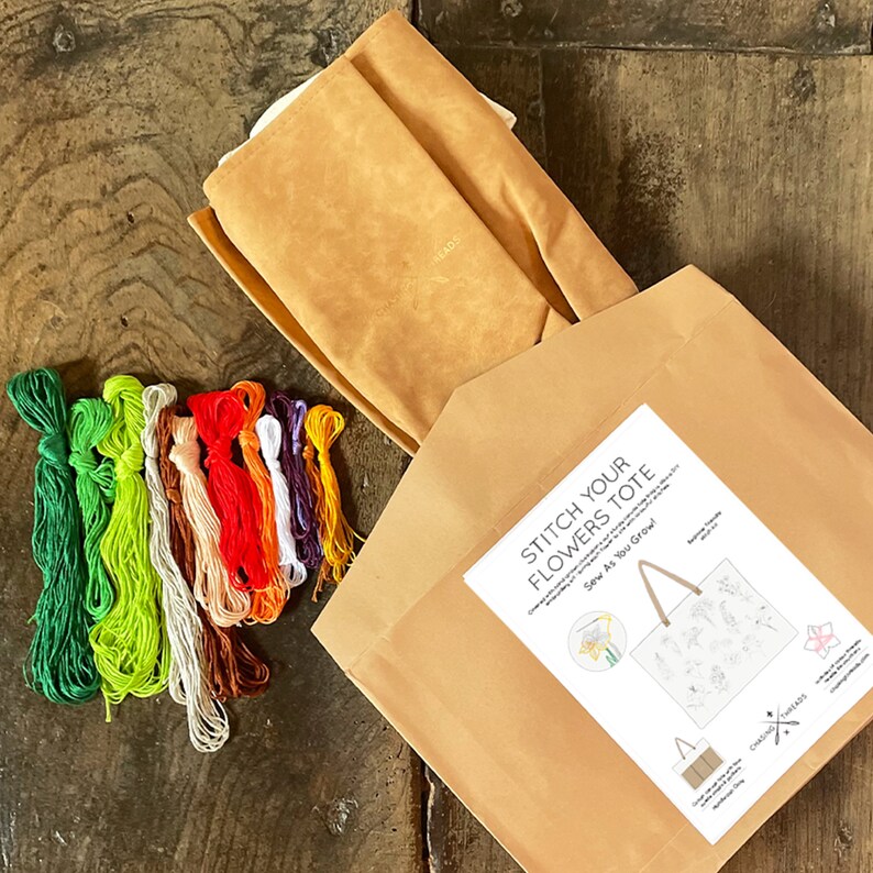 May include: A craft kit for a tote bag embroidery project. The kit includes a brown canvas tote bag, embroidery floss in various colors, and a pattern sheet with instructions. The pattern features a floral design with the text "Stitch Your Flowers Tote" and "Sew As You Grow!"