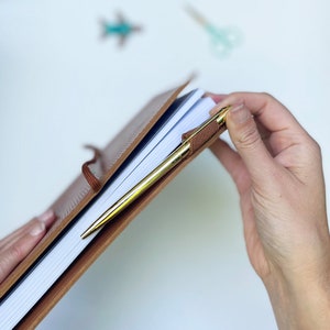 May include: A brown leather-bound journal with a gold pen tucked inside. The journal is open and the pages are white.