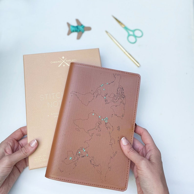 May include: A brown leather journal with a world map design. The map is outlined in black and has small green dots scattered across it. The journal is open and the cover is visible. The text "Stitch Not" is visible on the cover of the journal.