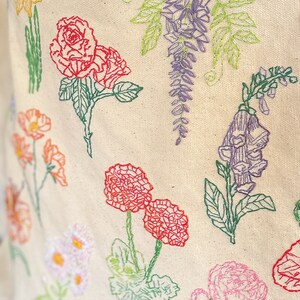 May include: A white fabric tote bag with embroidered flowers in various colors, including red, pink, purple, and green. The flowers include roses, carnations, and wisteria.