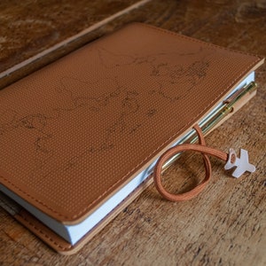 May include: A brown leather-bound journal with a world map embossed on the cover. The journal has a white page edge and a brown cord with a plane-shaped charm attached.