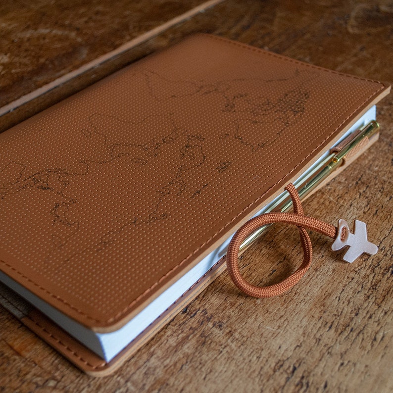 May include: A brown leather-bound journal with a world map embossed on the cover. The journal has a white page edge and a brown cord with a plane-shaped charm attached.