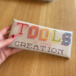 May include: A cream-coloured, rectangular case with the words "TOOLS OF MASS CREATION" embroidered in colourful cross-stitch. The text is in a gradient of colours, including red, orange, yellow, green, and blue. The case is held by a person.