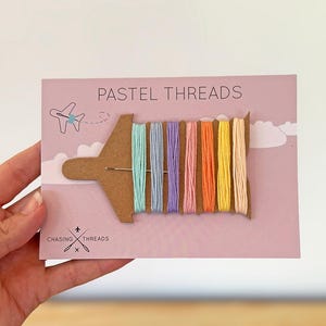 Può includere: Una scheda con fili pastello in vari colori, tra cui blu, rosa, arancione e giallo. La scheda presenta il testo "PASTEL THREADS" e un logo con la scritta "CHASING THREADS". I fili sono disposti su una forma di aeroplano di cartone.
