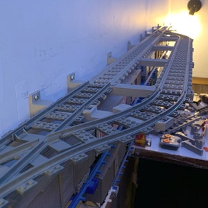 May include: A close-up of a grey Lego train track set up on a wall. The track is curved and has a points track. The track is mounted on a wall with brackets.